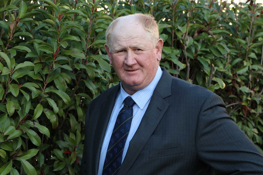 A middle-aged man in a suit and tie standing in front of some shrubbery.