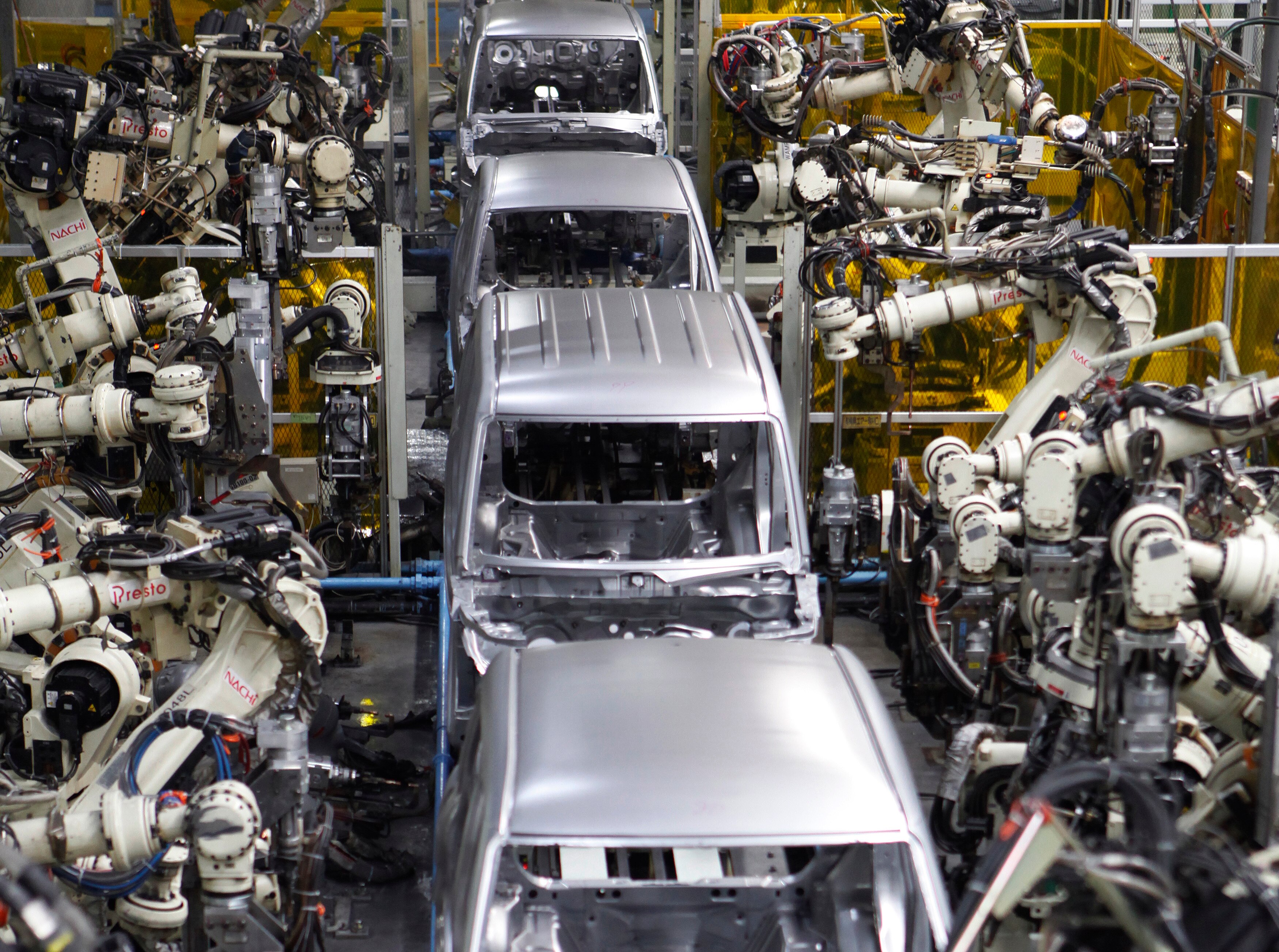 White, metal robotic arms work on a row of silver van bodies on a production line.