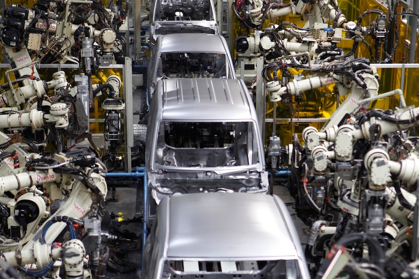 White, metal robotic arms work on a row of silver van bodies on a production line.