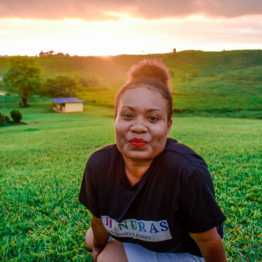 A woman in a black t-shirt sits smiling at the camera is a lush green field during sunset
