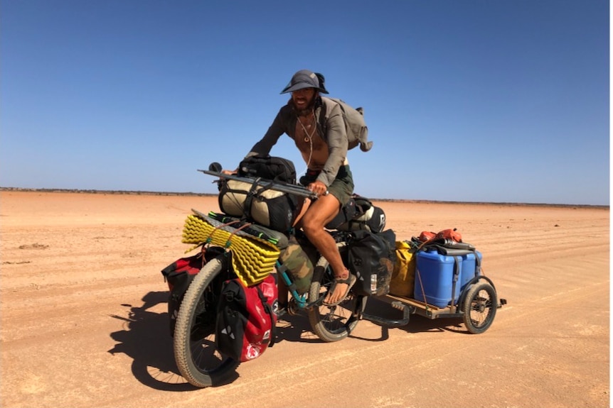 A man smiles atop a bicycle packed with camping gear on a dirt road in the desert.