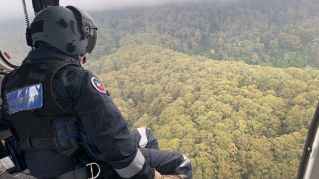 A man wearing a helmet leans out of an aircraft in cloudy conditions overlooking dense bushland.