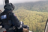 A man wearing a helmet leans out of an aircraft in cloudy conditions overlooking dense bushland.