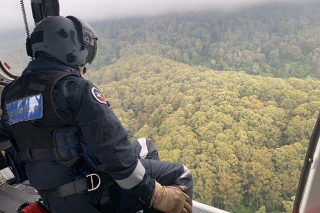 A man wearing a helmet leans out of an aircraft in cloudy conditions overlooking dense bushland.