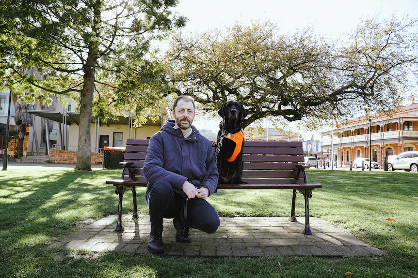 A man crouches beside a park bench with a black labrador sitting on it.