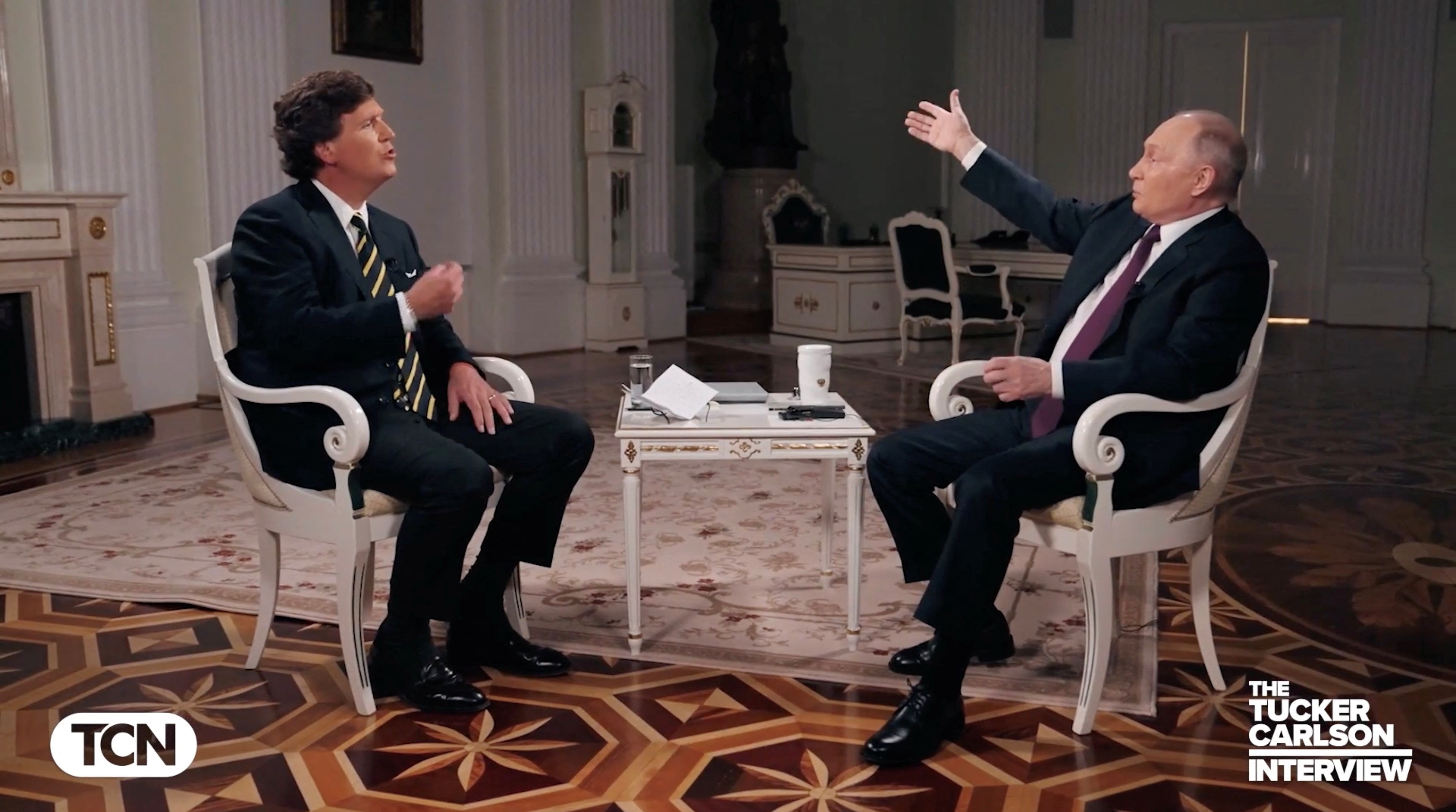 Vladimir Putin gestures while sitting in a chair across from Tucker Carlson