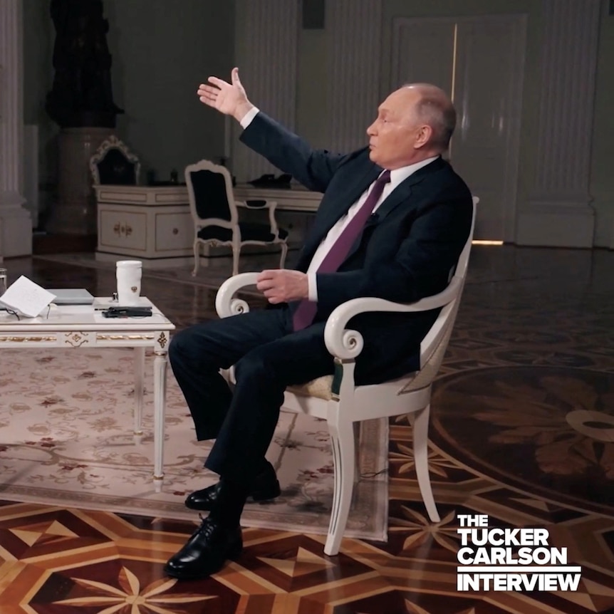Vladimir Putin gestures while sitting in a chair across from Tucker Carlson
