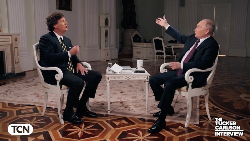 Vladimir Putin gestures while sitting in a chair across from Tucker Carlson