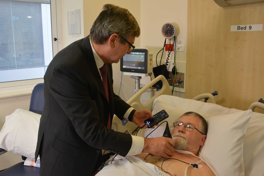 A man looks at a mobile phone screen while holding a device against a male patient's chest.