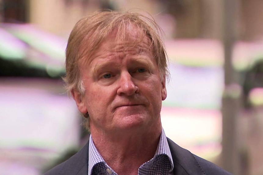 A man with fair hair wearing a suit jacket and shirt stands outdoors.