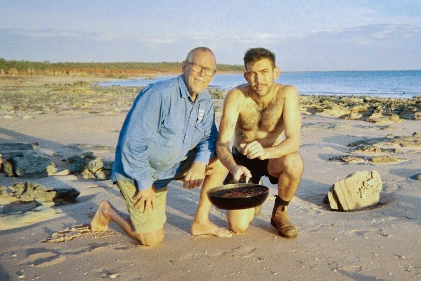 Two men kneel on a beach, Tom Forrest, right, holds out a frying pan to the camera. The image is lo-fi and grainy.