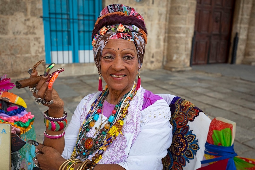 Une vieille femme cubaine avec de longs ongles colorés tient un cigare à la main et sourit, mettant en valeur un strass sur sa dent.