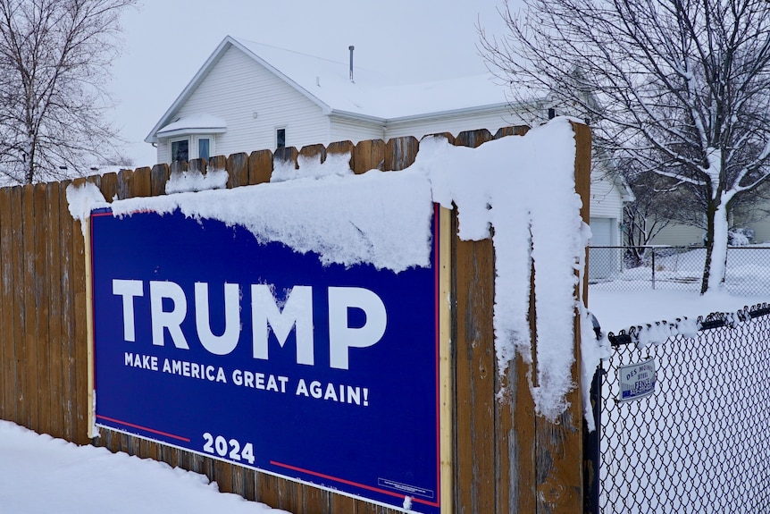 A sign on a fence in front of a snow-covered house says 'Trump Make America Great Again'