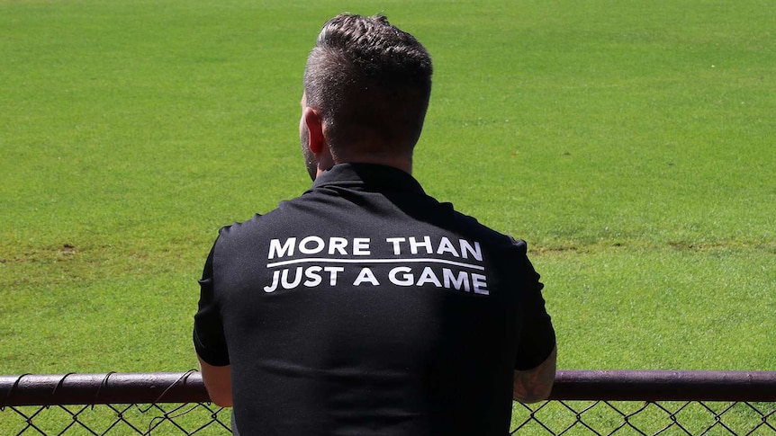 A man wearing a black shirt reading 'more than just a game' stands looking out over a football oval with his back to the camera.