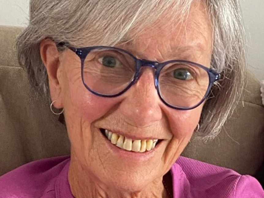 A close up of a woman's smiling face. She wears blue glasses and has a grey bob.