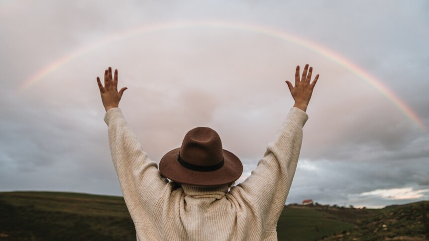 A person is standing with their back to the viewer, arms raised, looking at a rainbow in the sky