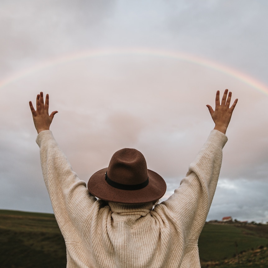 A person is standing with their back to the viewer, arms raised, looking at a rainbow in the sky