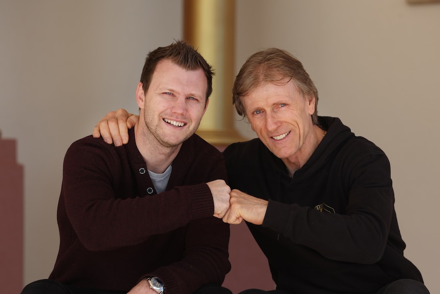 two men fist pump while sitting down, smiling at camera