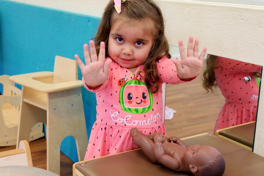 A little girl holds up her hands and smiles as she plays with a doll.