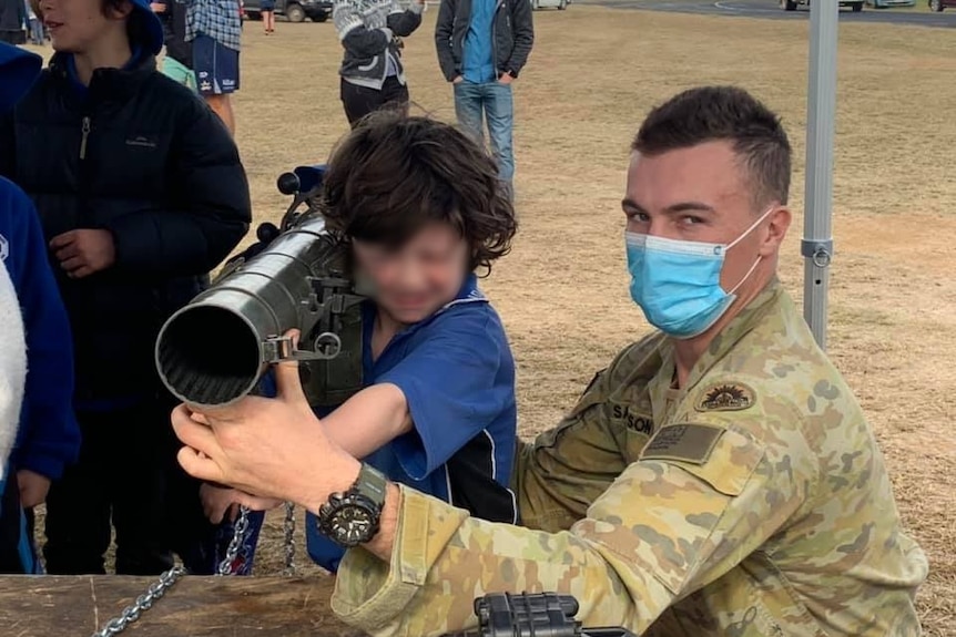 A small child in school uniform is almost dwarfed by an army weapon as a soldier in fatigues and a mask looks at camera