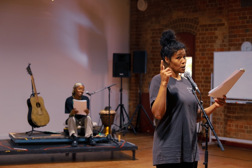 In a rehearsal room, Zahra Newman stands at a microphone, a script in hand, Kuda Mapeza seated behind.