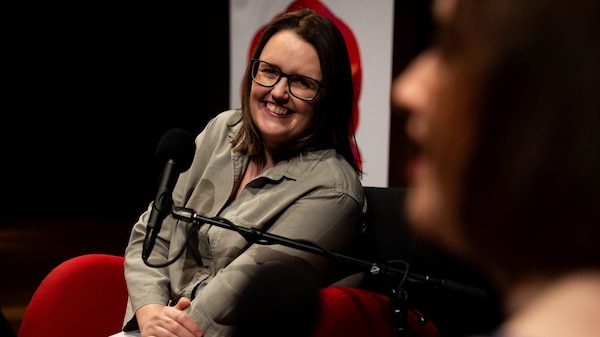 A smiling woman with brown hair and glasses sits in front of a microphone