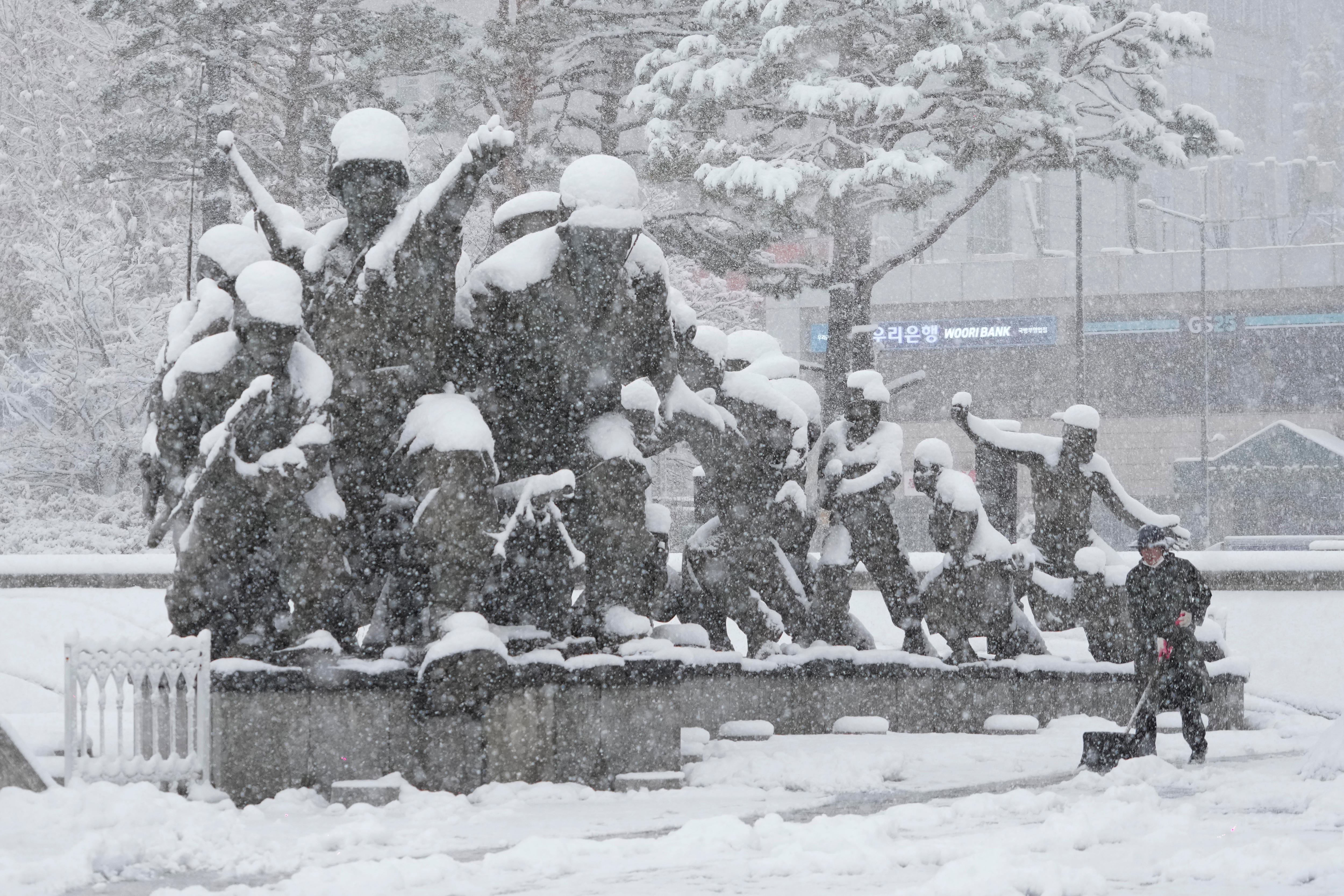 A worker wearing dark clothes using a shovel in deep snow in front of a statue of multiple soldiers