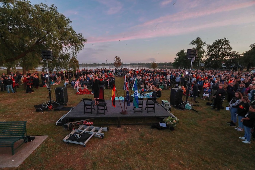 a podium, surrounded by a large crowd of people in the early morning light, with a lake  in the background.