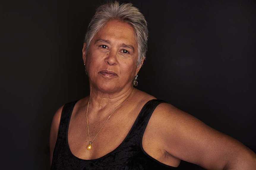 A woman with grey hair and brown skin wearing a black top looking at the camera.
