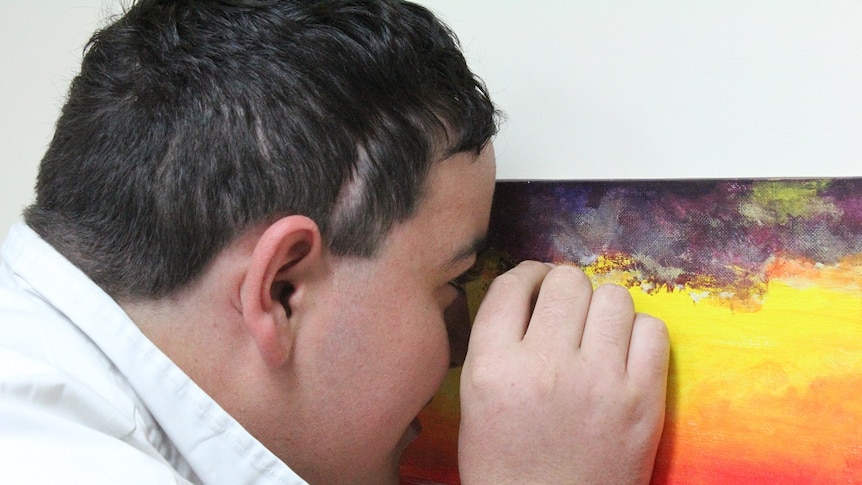 The side of a teen's face and head as he stands about a centimetre from the canvas, hand held up close as though holding a brush