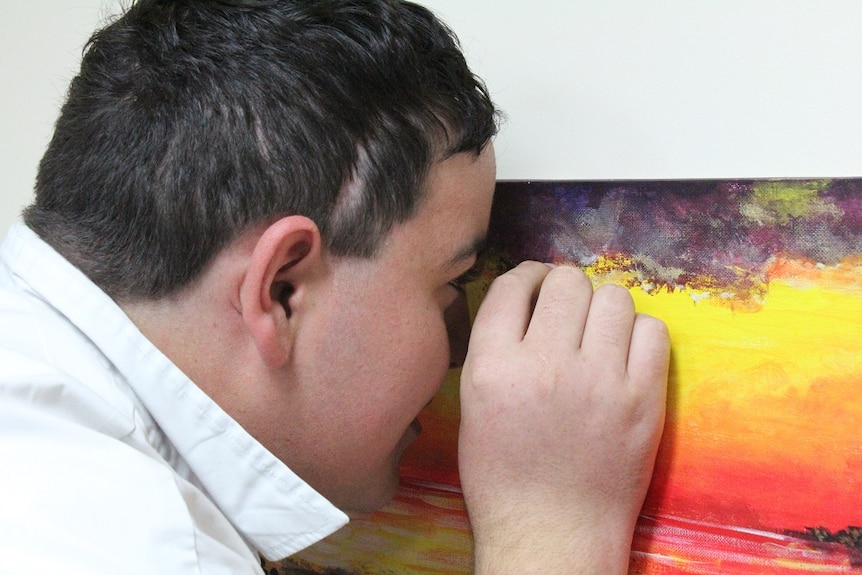 The side of a teen's face and head as he stands about a centimetre from the canvas, hand held up close as though holding a brush
