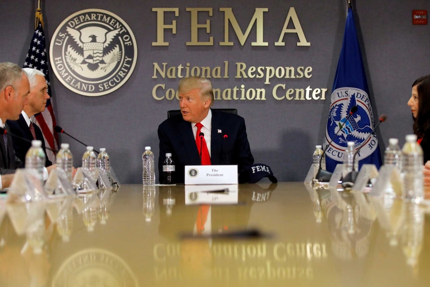 Donald Trump sitting at the top of a boardroom table with the US flag and the homeland security seal behind him.