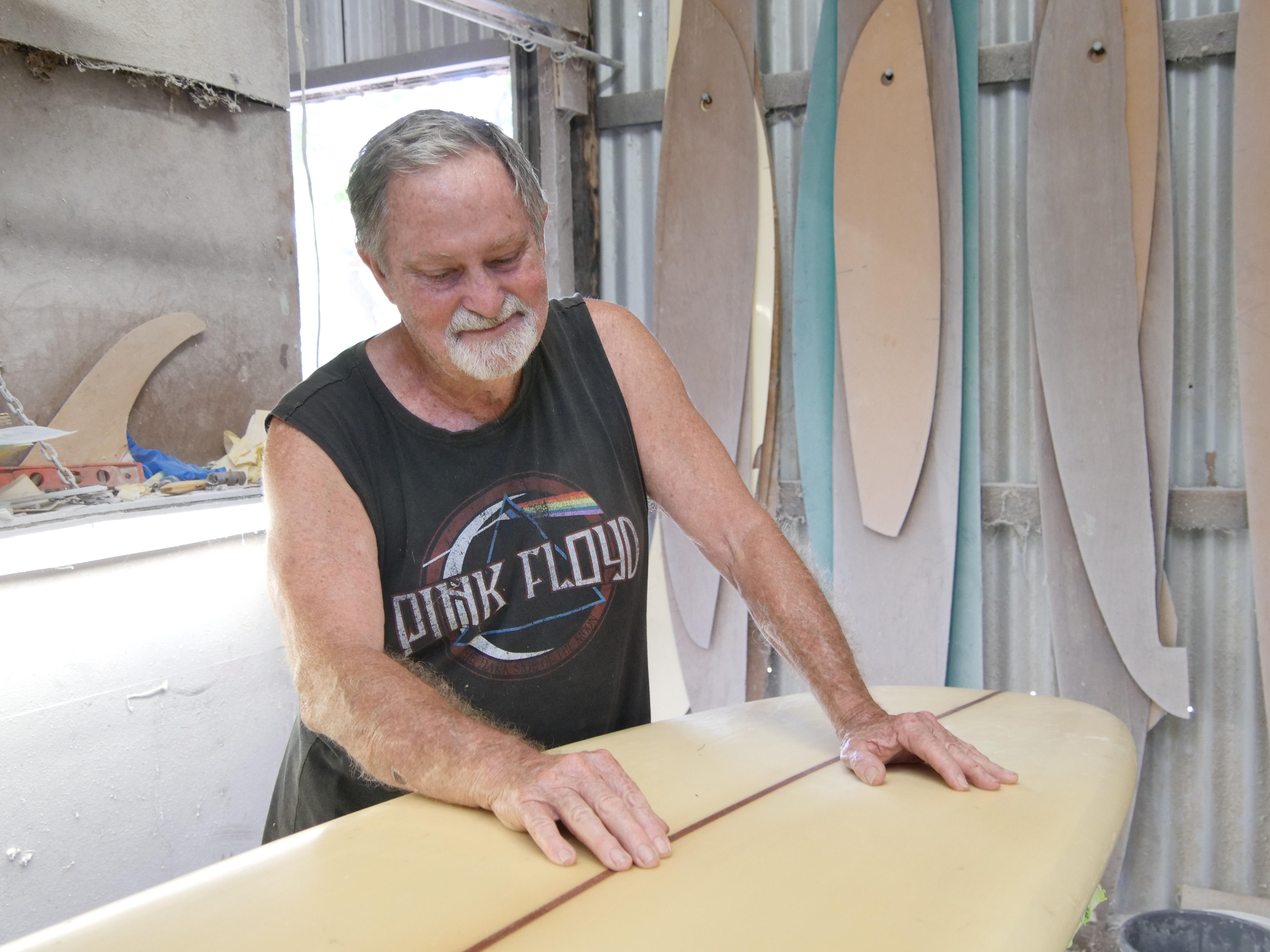 Hombre mirando una tabla de surf sobre una mesa en un cobertizo de trabajo con plantillas de tablas de surf al fondo.
