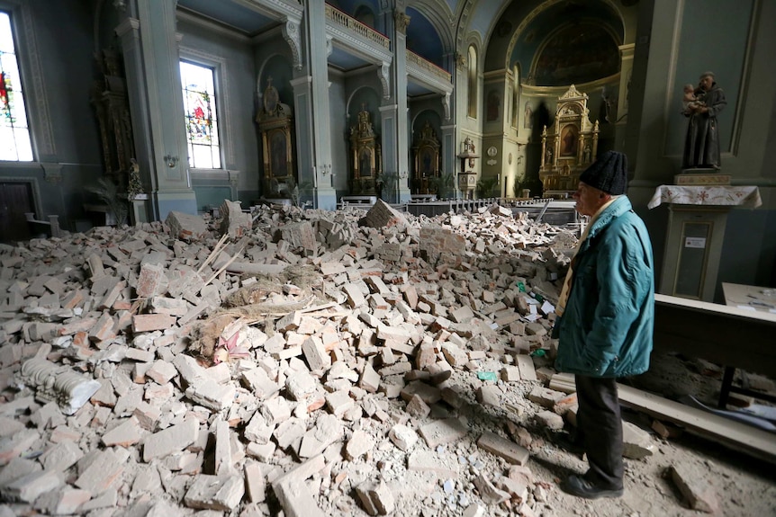 You see a damaged blue neoclassical church interior as an old man looks to a pile of debris on its floor.