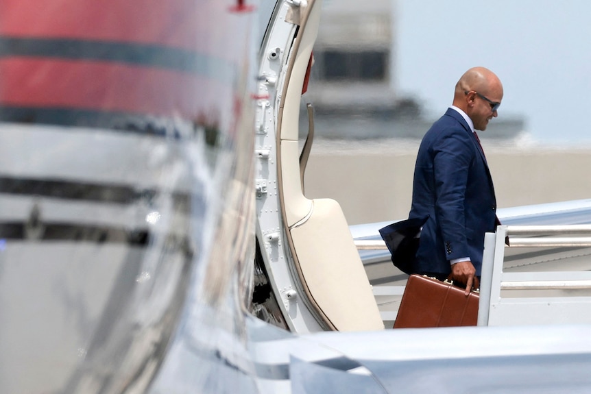 A bald man wearing a suit and carrying a briefcase steps off a plane onto stairs leading to the tarmac