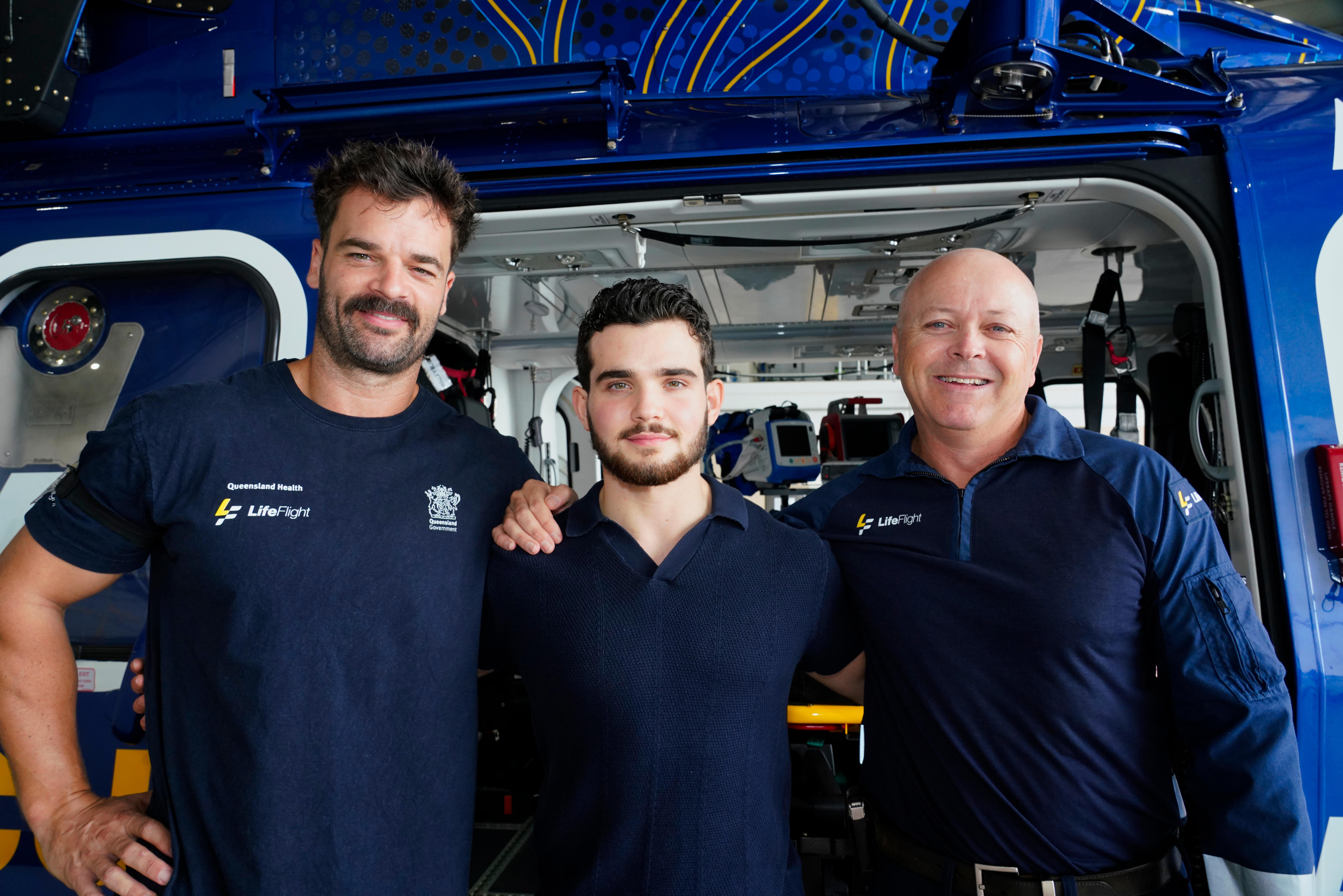 Dois homens com uniformes da LifeFlight estão um de cada lado de um adolescente de cabelos escuros na frente de uma ambulância.