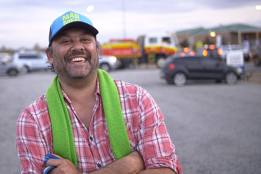 A man in a carpark smiles at the camera with his arms folded