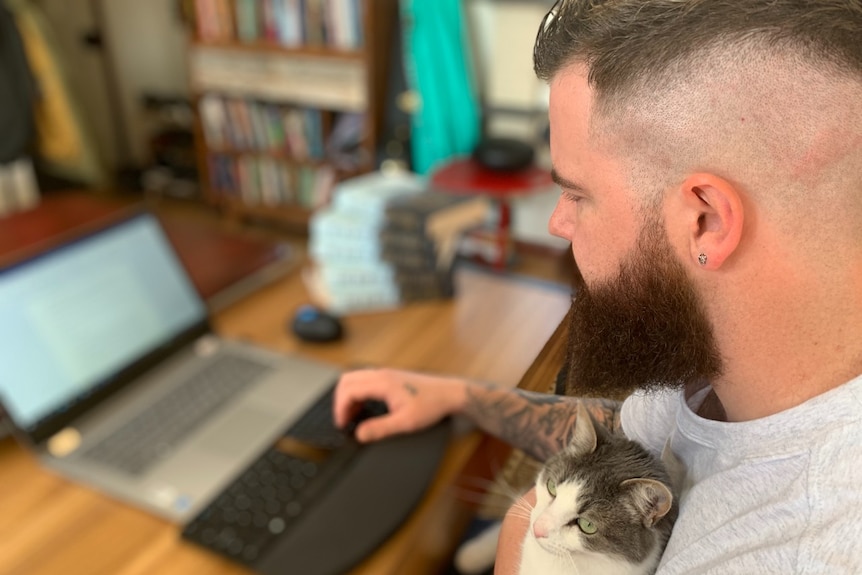 A man cradles a cat while using a laptop