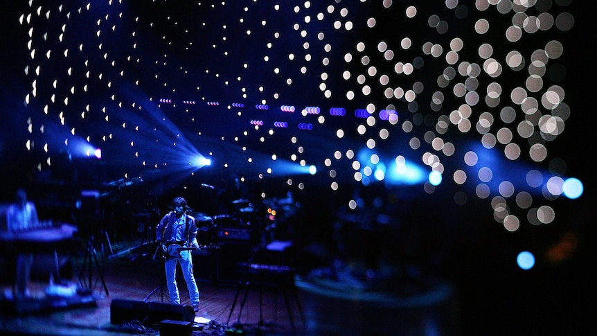 Nicholas Godin stands in on stage holding a guitar in the bottom corner of the image bathed in blue light and reflections