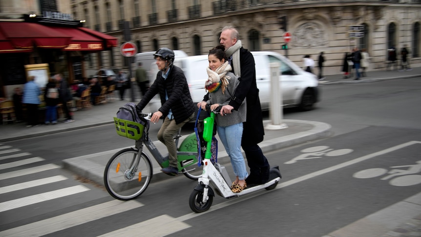 A man and a woman stand on a green-and-white e-scooter with no helmets, on a road in Paris.