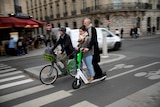 A man and a woman stand on a green-and-white e-scooter with no helmets, on a road in Paris.
