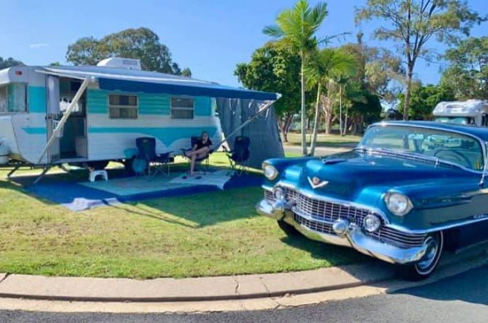 A man sits outside an aqua and white vintage caravan with a blue Cadilllac parked outside.