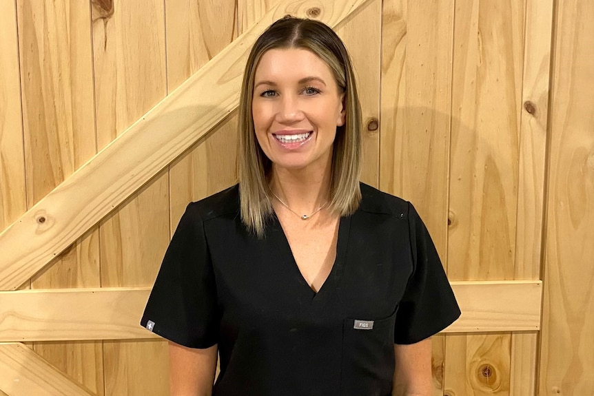 Woman with mid length hair, black t-shirt, smiling, stands in front of a wooden backdrop