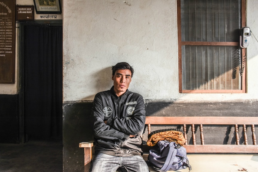 A man sits against a wall with his arms crossed while looking at the camera.