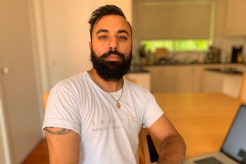 A man with short hair and a beard wears a white t shirt as he looks at the camera.