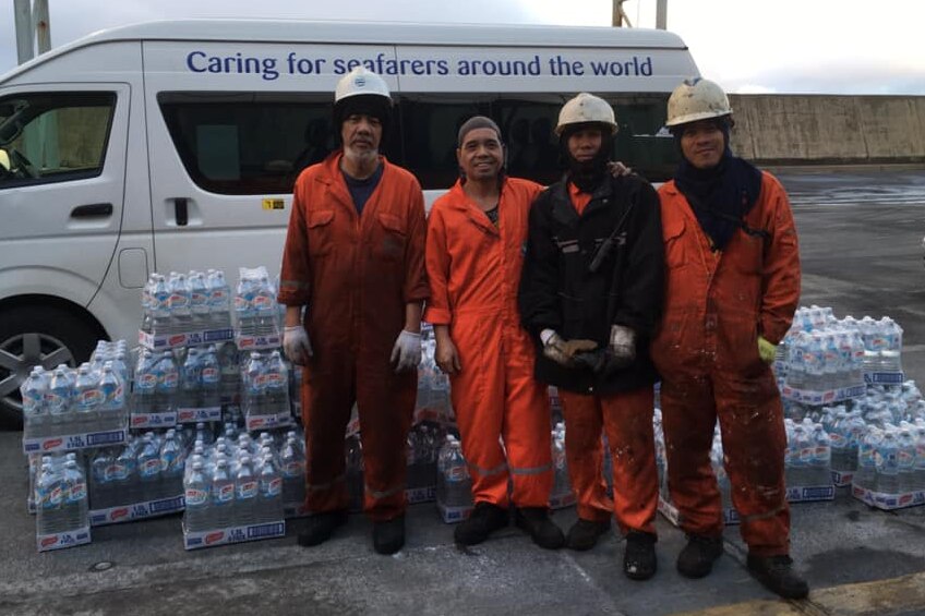 Four men in overalls and hard hats stand in front of piles of bottled water.