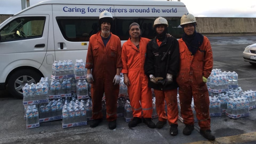 Four men in overalls and hard hats stand in front of piles of bottled water.