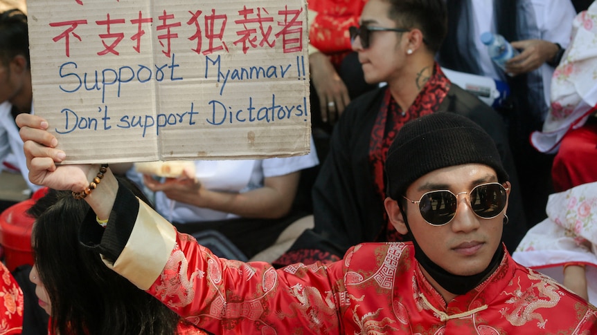 A man in a red traditional Chinese outfit holds up a sign at a protests that reads 'Support Myanmar, don't support Dictators'.