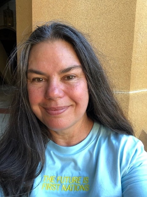 A portrait of a Indigenous Australian woman with long brown hair standing in front of artwork