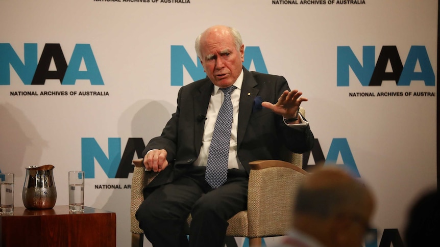 Former Prime Minister John Howard wears a dark suit with blue tie while sitting in a chair while gesturing with hand.